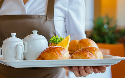 waitress in uniform serving breakfast with tea and pastries on tray in restaurant, fresh buns, elegant teapot, professional service, indoor dining background, focus on food Professional waitress presenting delicious breakfast on silver tray with teapot, cup, and freshly baked pastries in well-lit restaurant setting, hospitality concept